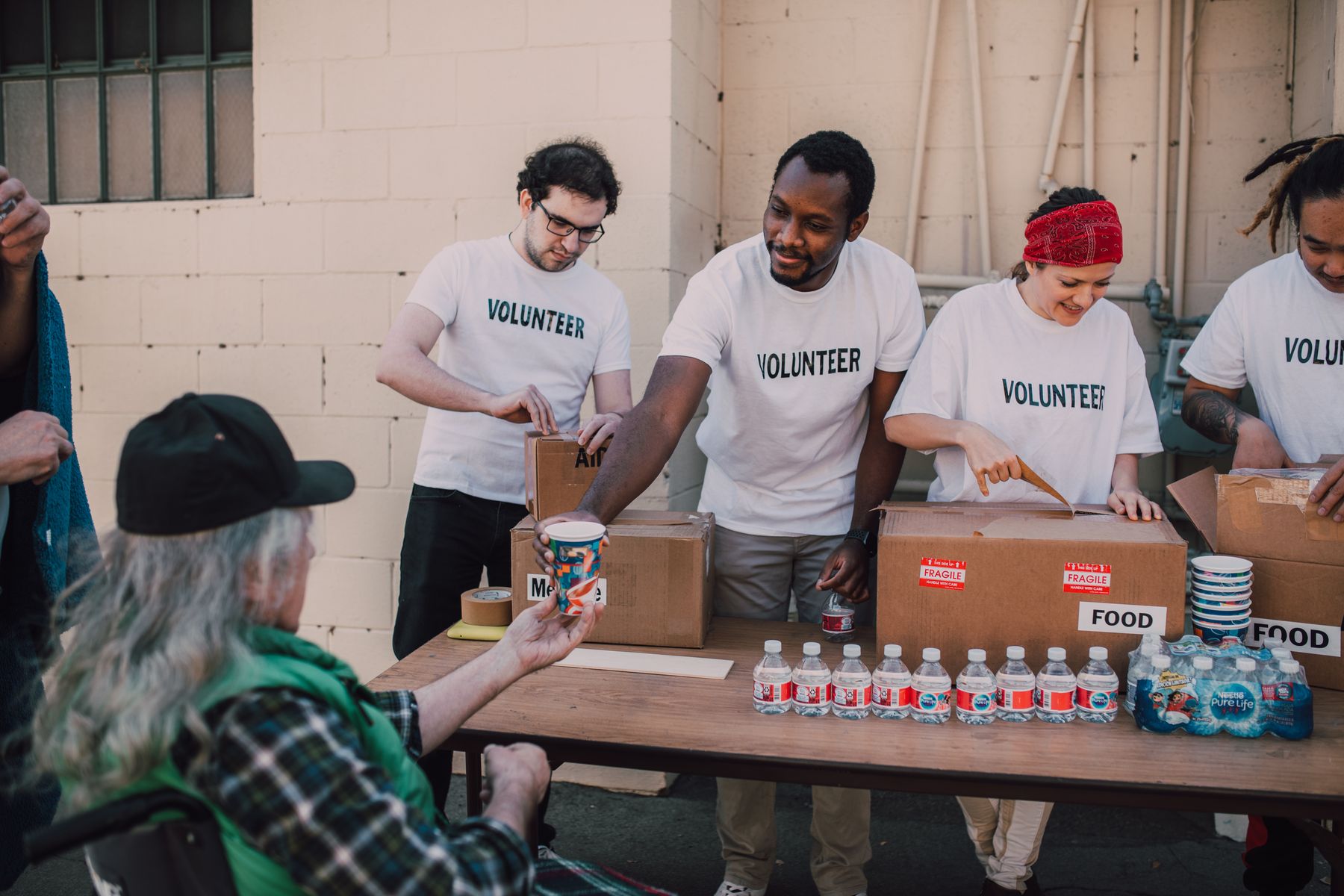 Volunteers at a folding table outdoors, handing a cup of coffee across to a man in a green jacket.