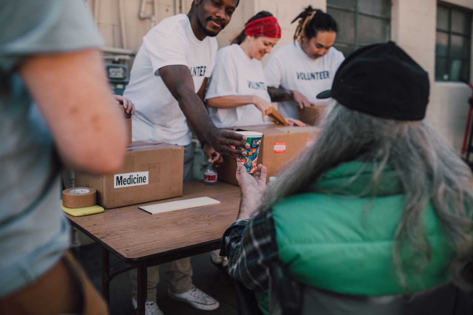 A volunteer reaching across a table to pass a cup to a seated man, boxes of food and medicine between them.