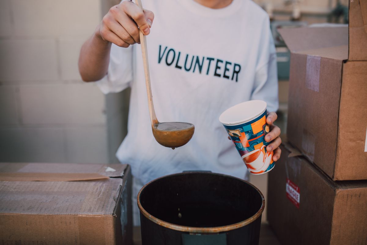 A volunteer in a printed T-shirt ladling soup into a paper cup.