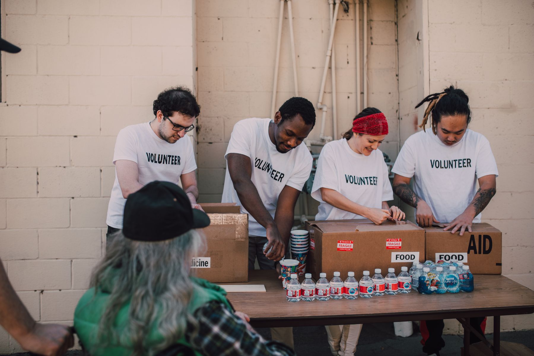 Four volunteers packing boxes of food and aid at an outdoor table, a man in a green jacket waiting in the foreground.